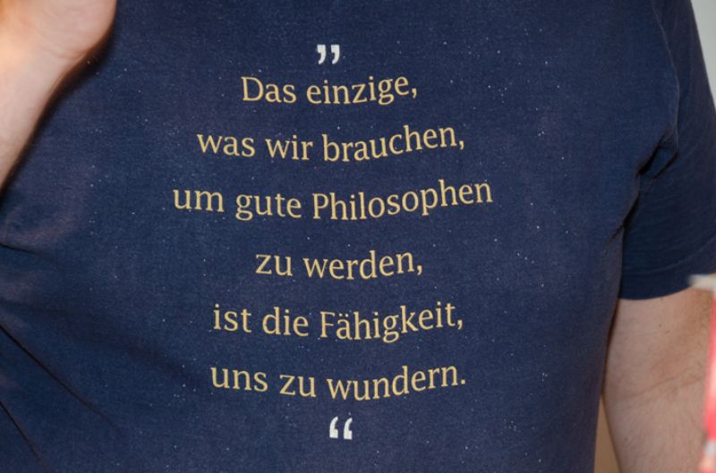 Een T-shirt met een filosofische uitspraak in het Duits: 'Das einzige, was wir brauchen, um gute Philosophen zu werden, ist die Fähigkeit, uns zu wundern.